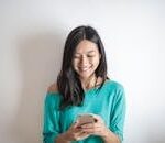 A happy woman in a green top smiling while using her smartphone indoors.