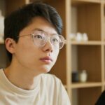 Portrait of a young man wearing eyeglasses, seated indoors with a blurred background.